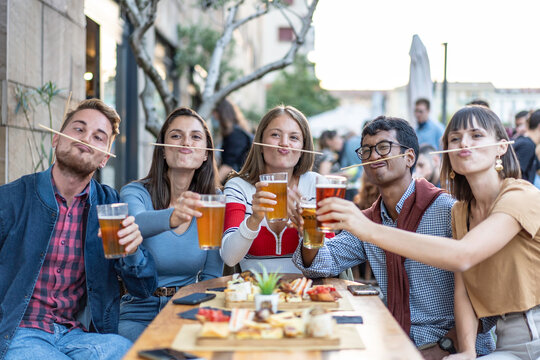 Playful friends toasting beer with chopsticks like mustaches at an outdoor cafe. Multiethnic millennials and gen z make silly faces during happy hour. Humor concept
