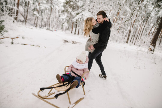 A father holding the mother in his arms while their baby sits in a sled, family winter walk in the forest. Closeup.