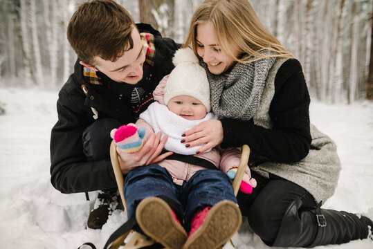 Happy parents sitting next to their baby who is in a sled, family winter walk in the forest. Dad, mom hugging their child sitting in a sled. Closeup.