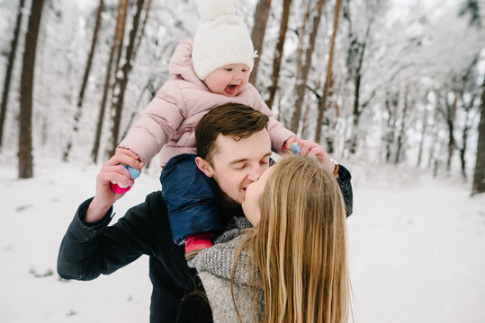 Couple kissing in snowy forest, father carrying happy little daughter on shoulders. Winter family vacation and family love. Closeup.