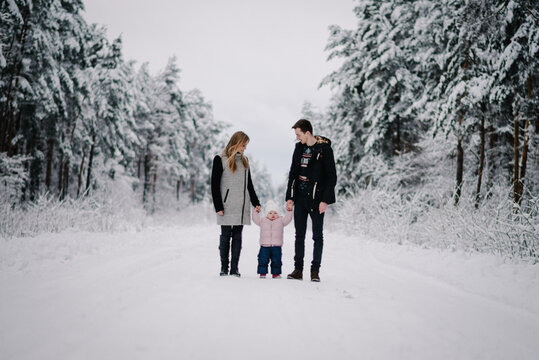 Happy family with child walking along snowy road in middle of winter forest. Dad, mom holding daughter by hands, enjoying joint walk. Winter holidays, comfort and joy in nature.