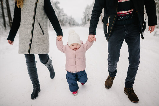 Happy parents holding hands with their little daughter learning to walk during a winter walk in a snowy forest. Family outdoor leisure. Down view.