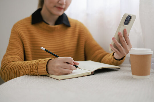 Young Asian Woman Studying with Smartphone and Notebook at Desk with Coffee Cup