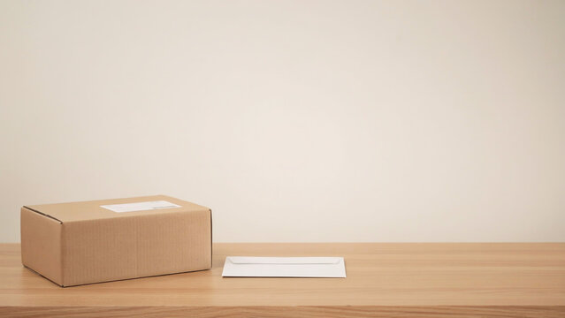 Small cardboard shipping box with label next to white envelope on light wooden table, representing mail, delivery, and correspondence services.