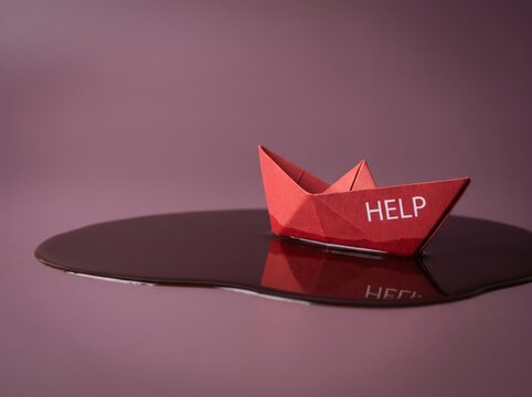 Red paper boat with HELP text on dark liquid puddle, distress signal