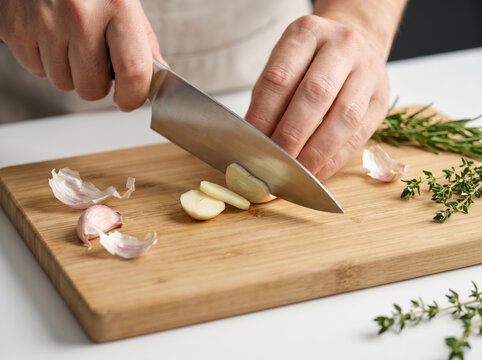 Chef hands slicing garlic cloves with a knife on a wooden cutting board
