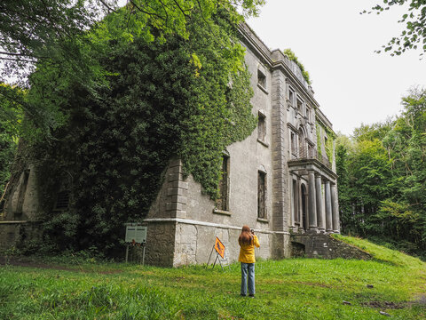 Girl tourist in yellow jacket explore ruins of stunning Moore Hall in county Mayo, Ireland. Huge old building covered with green vegetation in a public forest park. Travel and tourism in Ireland.