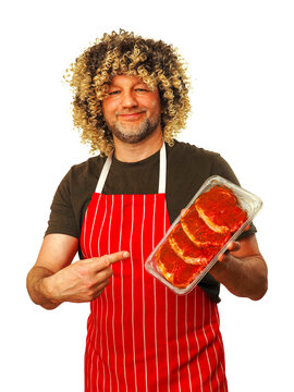 Man in his 40s or 50s with curly hair holds tray of marinated strip loin steaks. He wears red and white striped apron. Background is plain white. Funny looking butcher promoting his craft product.