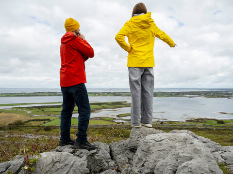Father in red coat and daughter in yellow travel jacket are on top of a rough stone in a mountains, stunning nature scene with ocean and cloudy sky in the background. Divorced family concept. Travel.