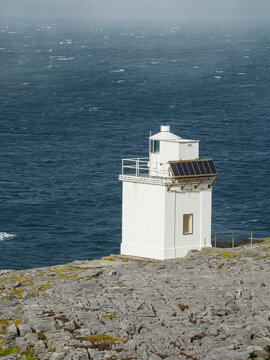 White Black head lighthouse stands on rough stone coast. Burren, Ireland. Ocean stretches wide, meeting sky with clouds. Rocky terrain shows nature raw, untamed strength. Tourist attraction.