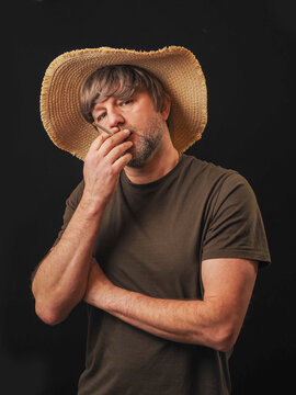 Man in his 40s or 50s wearing green shirt and big straw hat, dark background. Country looking male model with grey beard and friendly face. Holding cigarette in his hand. Bad boy theme.