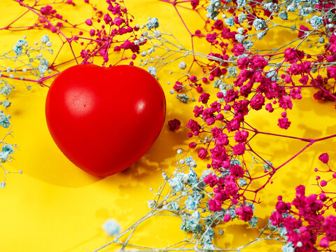 Vibrant red heart sits on bright yellow background. Surrounding flowers include pink and light blue baby breath. Delicate stems frame the central heart shape. Floral arrangement creates romantic mood