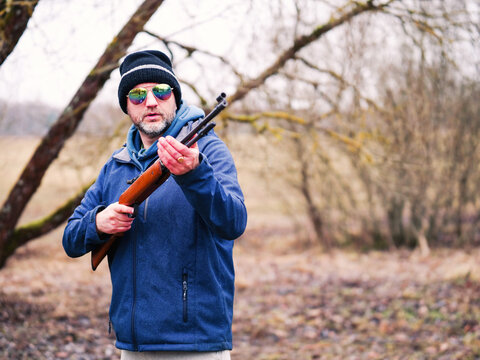 Person aims rifle outdoors in wooded area. Bare branches frame the scene. Ground covered with fallen leaves. Season appears late fall or early winter. No animals or plants identified