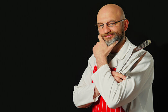 Bald man with glasses and beard stands confidently. He wears white coat and red striped apron. Holds spatula in hand. Smiles at camera. Black background highlights his figure