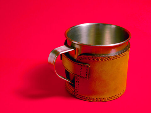 Stainless steel cup with leather wrap and handle. Red background highlights cup texture. Simple, modern design for outdoor or rustic use