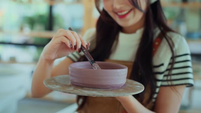 Beautiful young Asian woman painting ceramic bowl with brush in pottery studio, creative female artist working on handmade craft, pottery art and lifestyle concept