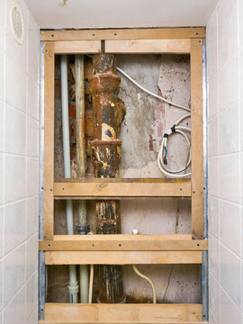 Exposed plumbing and wooden framing in tiled bathroom wall. Rusty pipes and exposed fixtures show renovation progress. Brown water filter attached to wall below frame