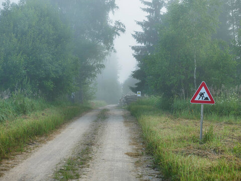 Misty forest path with construction warning sign. Gravel road winds through dense green trees. Fog partially obscures distant trees. Sign shows worker silhouette. Woodcutting area.