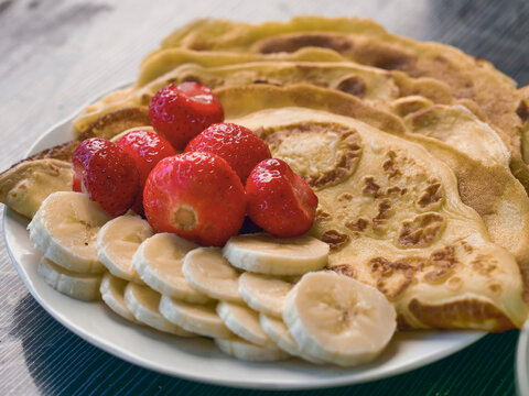Stack of golden-brown pancakes on white plate. Fresh strawberries and sliced bananas beside pancakes. Soft and dreamy look, Hand made simple country meal. Dark wooden surface background