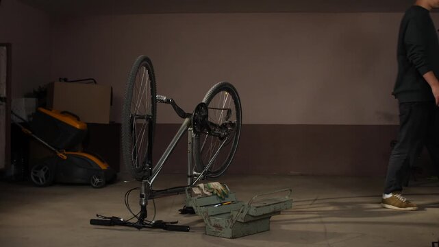 Man walking past an upright bicycle being repaired, with a toolbox on the floor and a lawnmower in the background of a domestic workshop