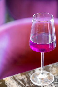 A glass of vibrant purple cocktail or wine sits on a dark marble tabletop, with a soft, blurred magenta background