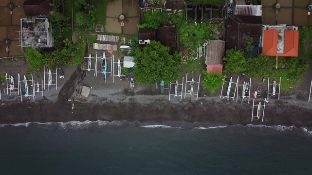 Traditional outrigger canoes rest on a narrow rocky beach beside dense trees and village houses along the Bali coastline, seen from directly overhead.
