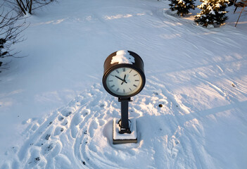 Naklejka premium Outdoor clock in a snowy landscape with footprints and evergreen trees, winter season and time concept, an outdoor clock stands in a snowy landscape