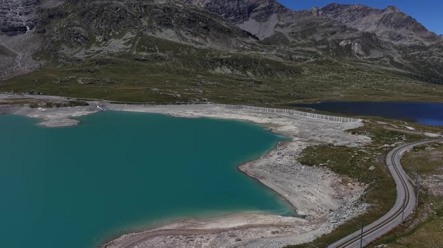Aerial view of the turquoise waters of Lago Bianco reservoir and the winding Bernina Pass railway tracks set against rugged alpine mountains in Poschiavo, Grisons, Switzerland.