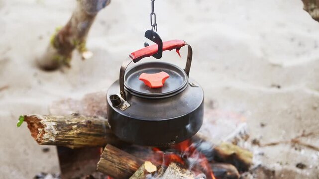 Outdoor campfire kettle hanging over glowing bonfire logs with sandy background natural setting
