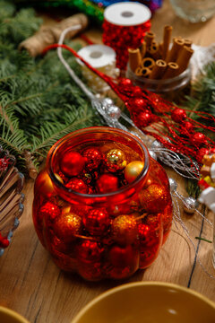 Vertical still life of red glass jar filled with shiny red and gold christmas baubles surrounded by pine branches cinnamon sticks ribbons and glitter garlands on wooden table