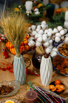 Vertical still life of dried wheat and cotton branches in geometric vases surrounded by tangerines candles glitter wires and spices on wooden table for cozy holiday decor