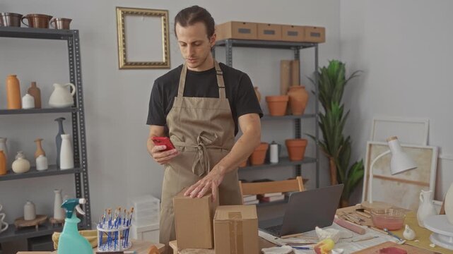 Man in apron holds phone and places cardboard box with bare hands in studio workspace; small business focused.