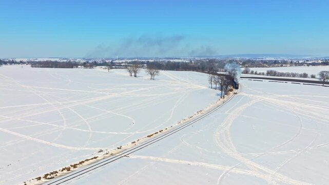 A steam train travels along tracks in a snowy field. Smoke rises as the train moves. The bright blue sky contrasts with the white snow. The scene shows a winter day.
