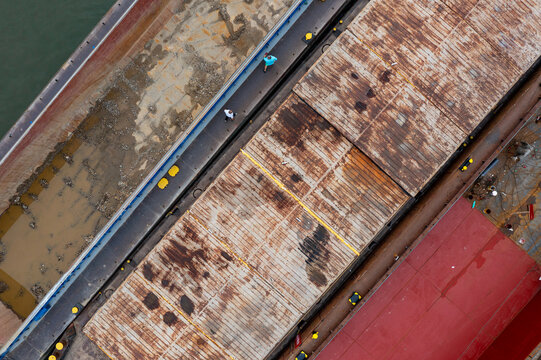 Aerial view of a large industrial barge with a rusted metal deck and a red section moored on the Danube River, with workers walking along the side walkway in Pancevo, Vojvodina, Serbia.
