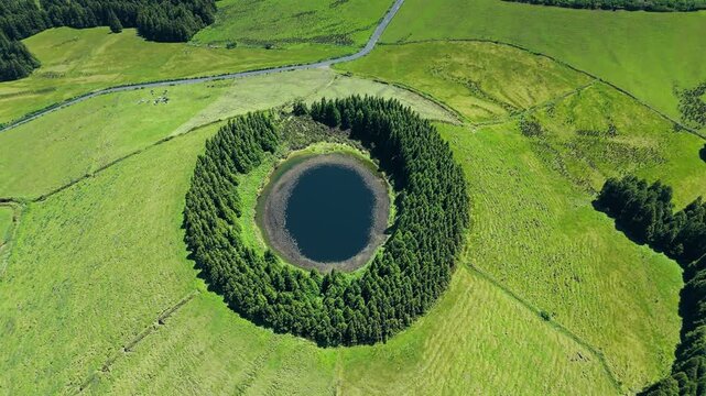 Volcanic lake with dark reflective surface bordered by circular forest on Sao Miguel Island, drone footage