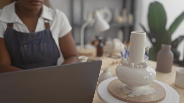 Woman potter in denim apron with hands on table, looking at laptop and glazed vase on wheel in studio; calm concentration.