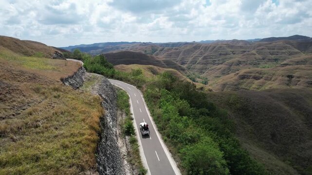 Aerial drone footage of a winding road crossing Sumba&rsquo;s rolling hills, with a vehicle, dramatic valleys, dry grasslands, lush trees, and cloudy blue sky creating a scenic travel landscape.