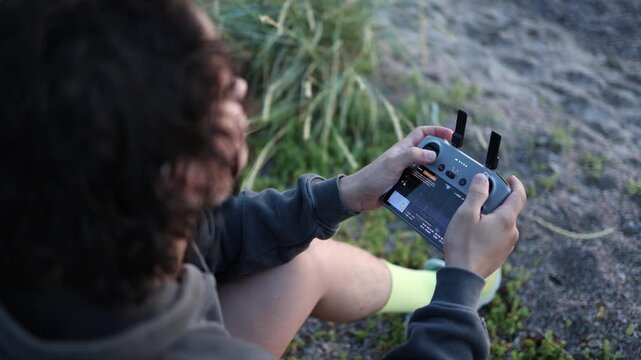 Young curly haired man controlling a drone with a remote controller in his hands while focusing on the flight and operating the joysticks outdoors