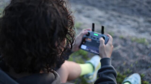 Curly haired young man holding a drone remote controller and operating the control sticks while flying a drone outdoors in natural light