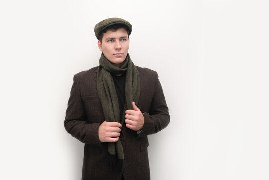 Young man posing in a brown winter coat with wool scarf and flat cap, offering a thoughtful, confident fashion image. The guy is very serious, sad, doesn't want to grow up, to be responsible