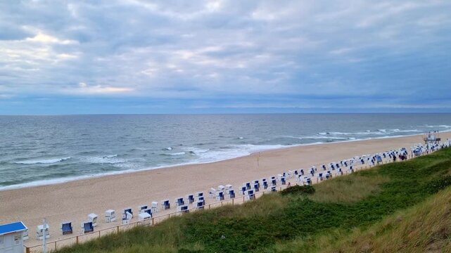 Coastal beach scene with rows of blue and white chairs along sandy shore, gentle waves lapping at the beach under a cloudy sky in a tranquil seaside setting