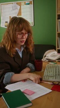 Vertical video of young adult Caucasian woman with glasses writing on document at desk, then opening red notebook to check notes. Clip is ideal for retro office or vintage business concepts