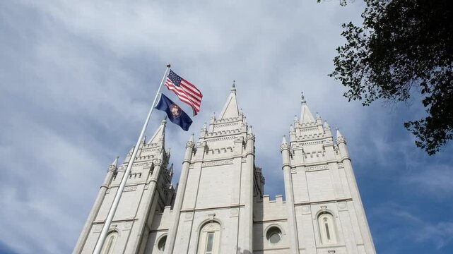 Exterior of the LDS Temple in Salt Lake City, Utah on Temple Square, bathed in sunlight, clouds gently rolling by overhead.  American and Utah State flag gently waving on a flag pole .