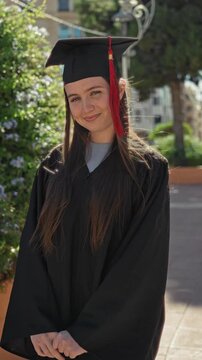 Woman in graduation gown and mortarboard with red tassel, smiling face and clasped hands outside building; achievement pride.