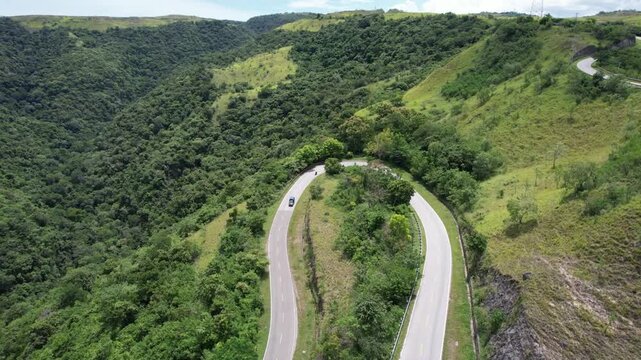 Cinematic view of a winding mountain road weaving through Sumba&rsquo;s lush green valleys, dense tropical forest, and rolling hills beneath bright daylight, capturing peaceful natural scenery.