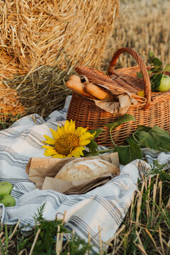 A woven basket filled with bread and apples sits beside a golden hay bale, with a sunflower and rustic blanket completing the countryside scene.