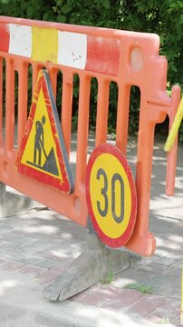 Orange construction barrier with warning sign and speed limit indicator positioned on a sidewalk, surrounded by greenery and pavement, indicating work zone ahead
