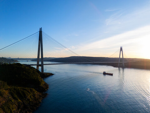 Aerial vertical view of Yavuz Sultan Selim Bridge at sunset in Istanbul, Turkey. Modern cable-stayed bridge over Bosphorus Strait with cargo ship, small boat, rocky coast and golden sun light.