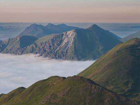 Aerial view of the rugged mountain peaks of Unalaska rising above a thick layer of low-hanging clouds under a soft evening sky in Unalaska, Alaska, United States.