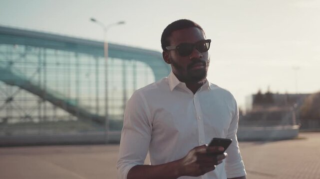 Handsome bearded man walks to the airport entrance and uses his smartphone, laughs at received message. Modern communication, being online, social networks, cheerful emotions. Male portrait